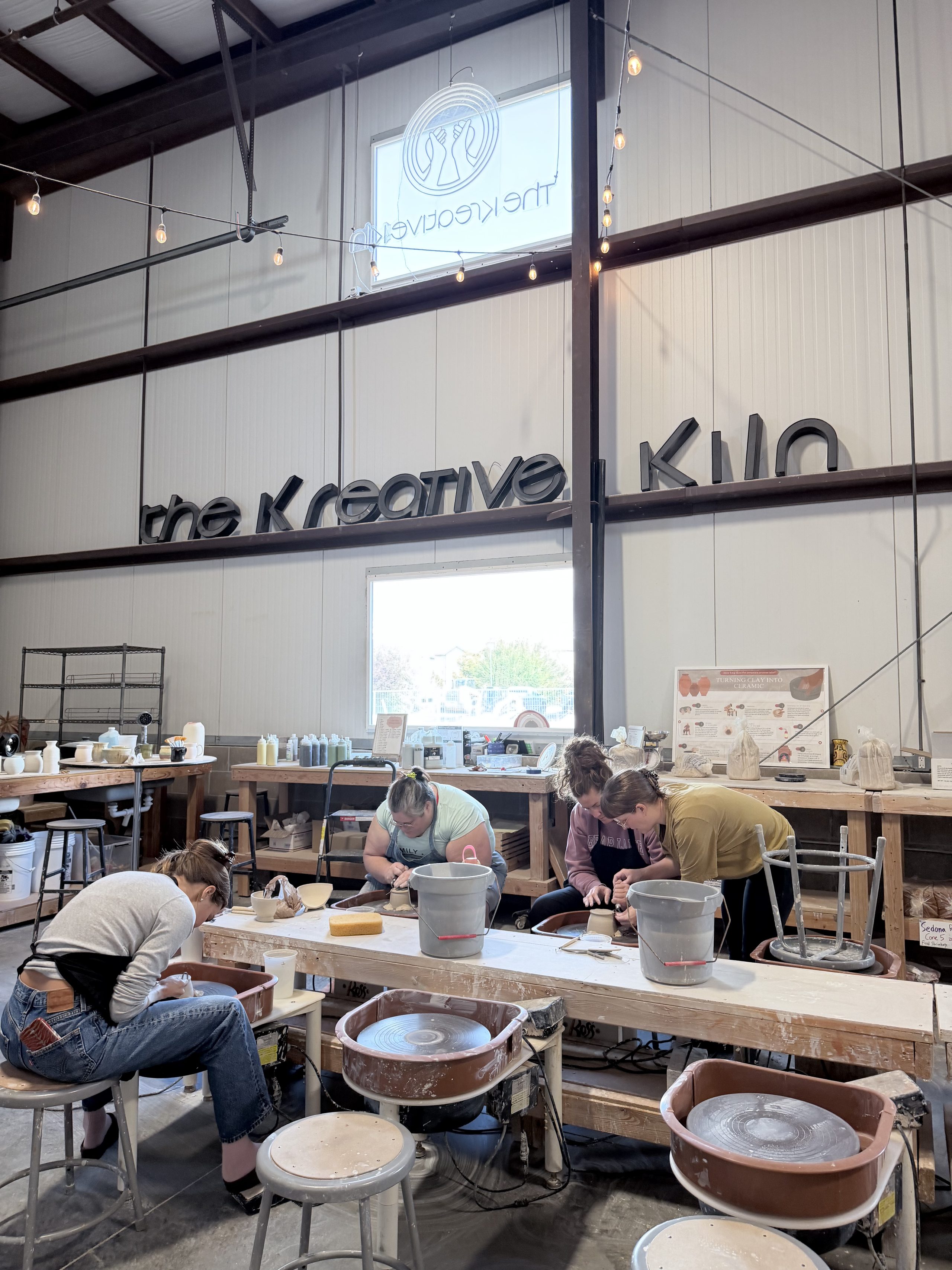 Students throwing at the wheels beneath the Kreative Kiln sign